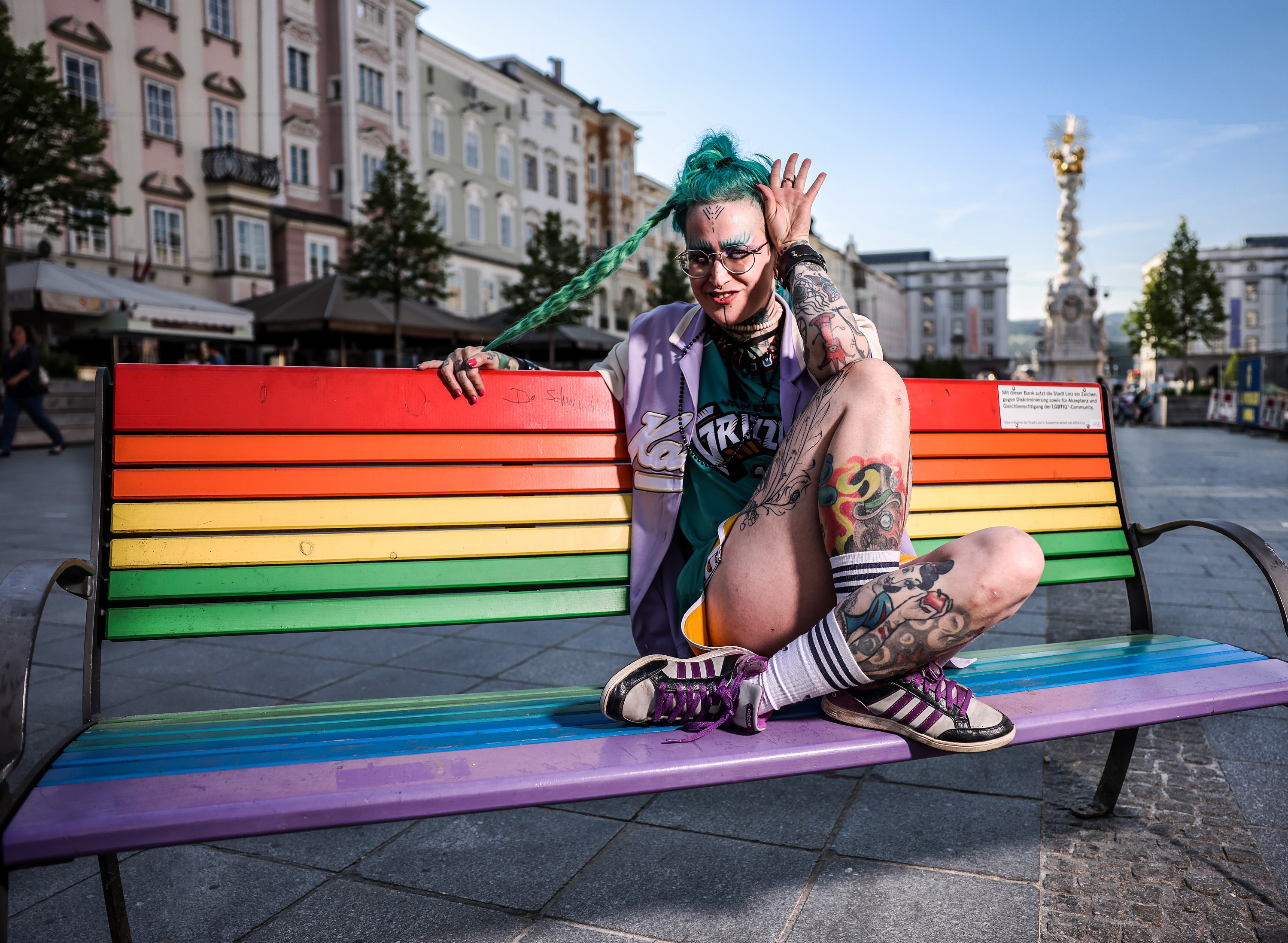 In den Farben des Regenbogens gestrichene Bänke zieren verschiedene Plätze in Linz als Zeichen für Toleranz. Hier sitzt Queer Artist Alice Moe auf einer der Bänke am Linzer Hauptplatz.